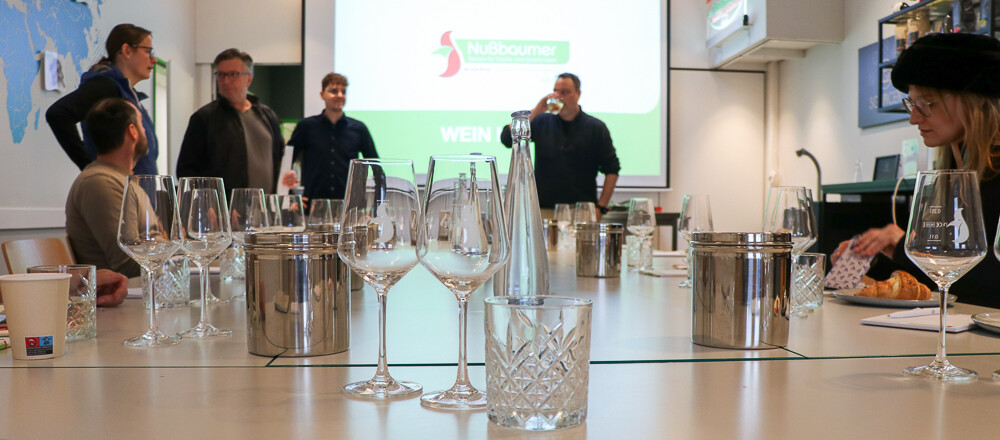 Eine Gruppe von Menschen steht um einen Tisch mit Gläsern und einer Projektionsleinwand.