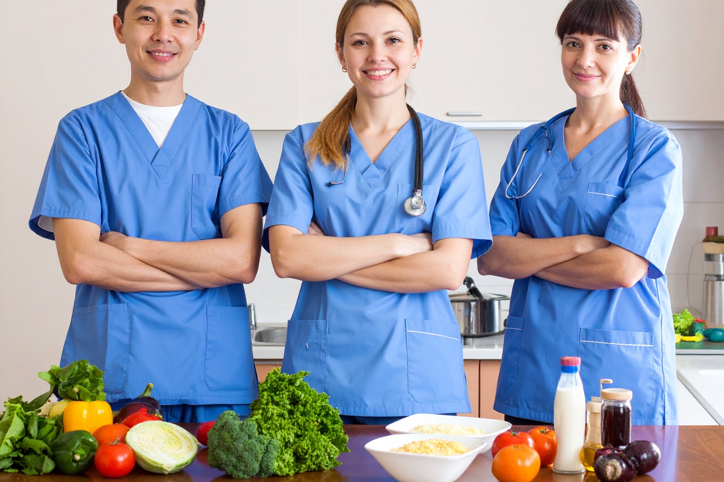 Eine Gruppe von Personen in blauen Kitteln steht lächelnd vor einem Tisch mit Lebensmitteln. Eine Gruppe von Personen in blauen Kitteln steht lächelnd vor einem Tisch mit Lebensmitteln.